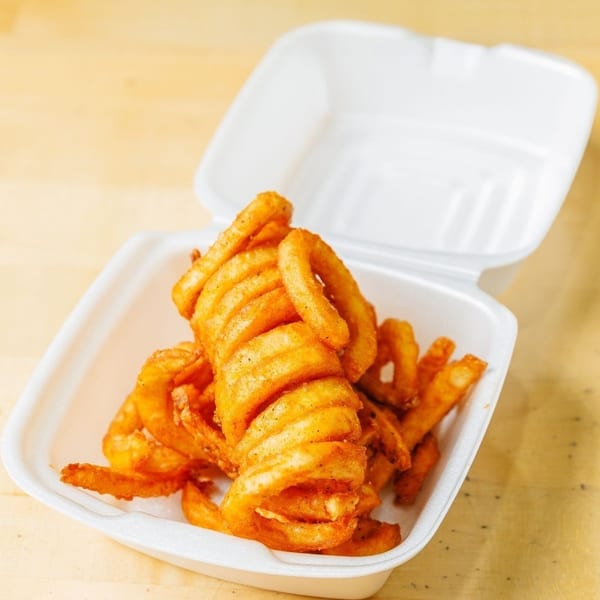 Seasoned curly fries in a white styrofoam takeout container on a light wooden surface.