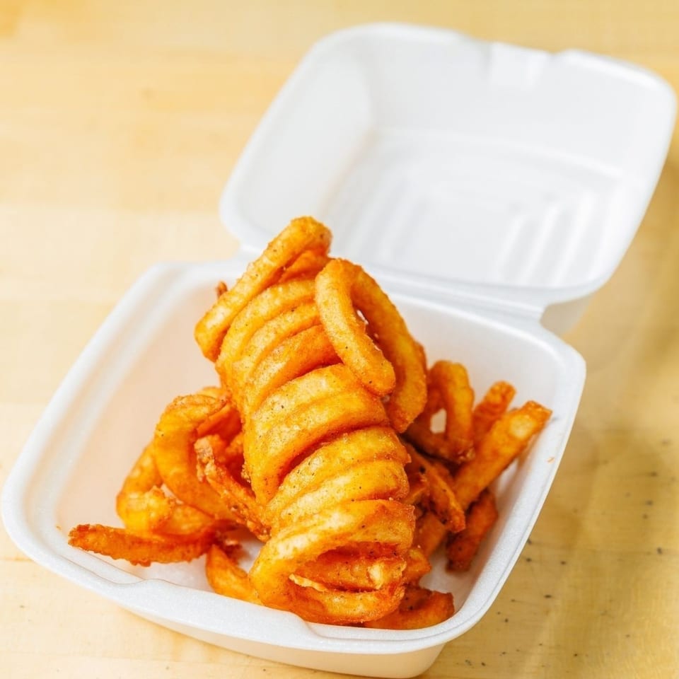 Seasoned curly fries in a white styrofoam takeout container on a light wooden surface.