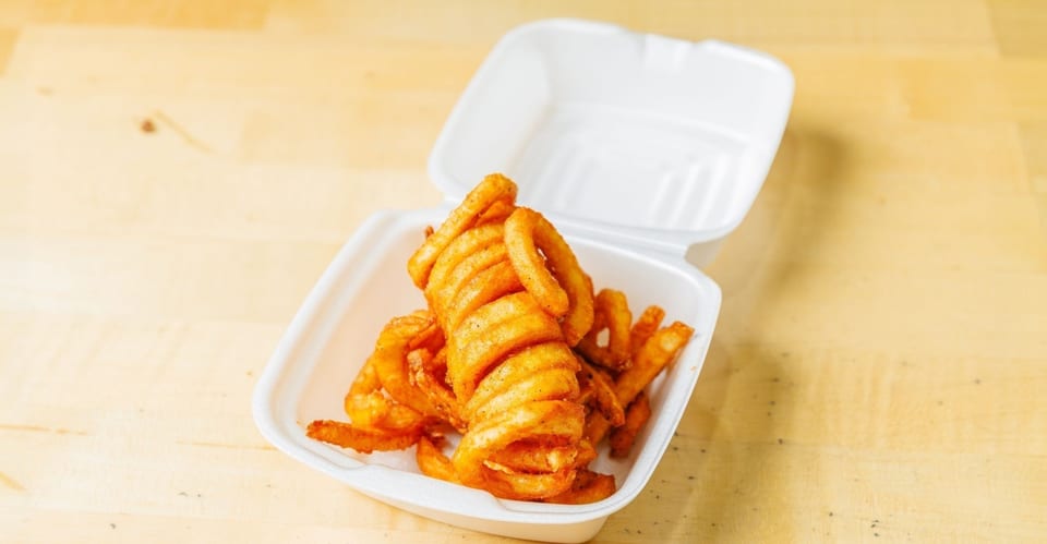 Seasoned curly fries in a white styrofoam takeout container on a light wooden surface.