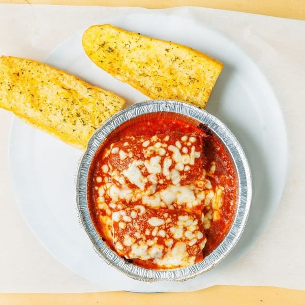 Baked ziti with melted cheese and tomato sauce in a foil container, served with two garlic breadsticks.