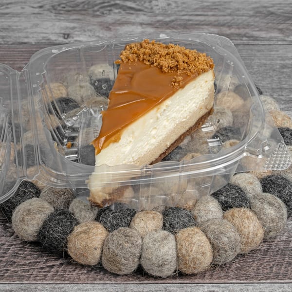 A slice of cheesecake with caramel topping and cookie crumbles in a plastic container.