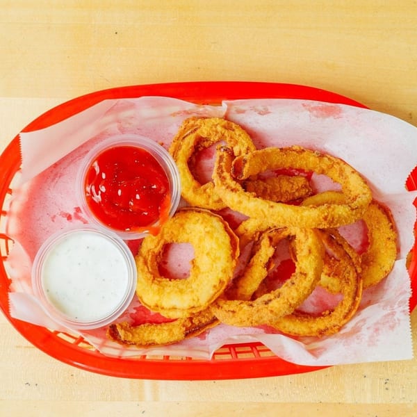 Golden-brown onion rings in a red basket with ketchup and ranch dipping sauces on a wooden table.
