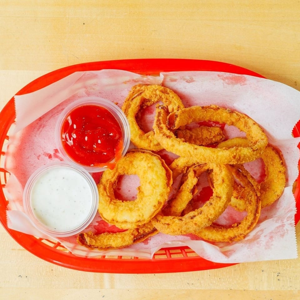 Golden-brown onion rings in a red basket with ketchup and ranch dipping sauces on a wooden table.