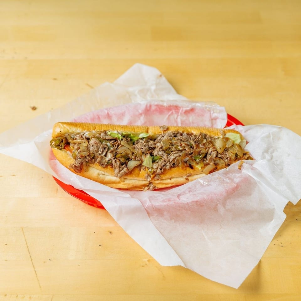 A large cheesesteak sandwich with steak, onions, and peppers in a red basket on a wooden table.