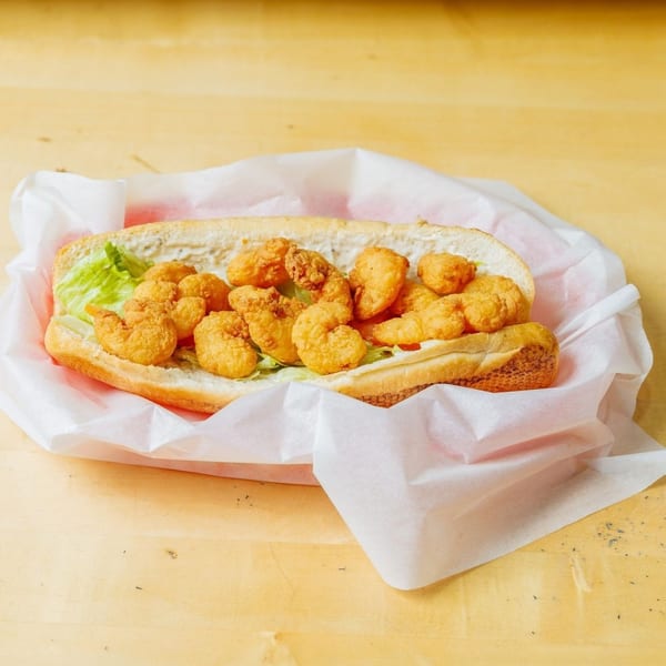 A shrimp po'boy sandwich with lettuce and sauce, wrapped in white paper on a wooden table.
