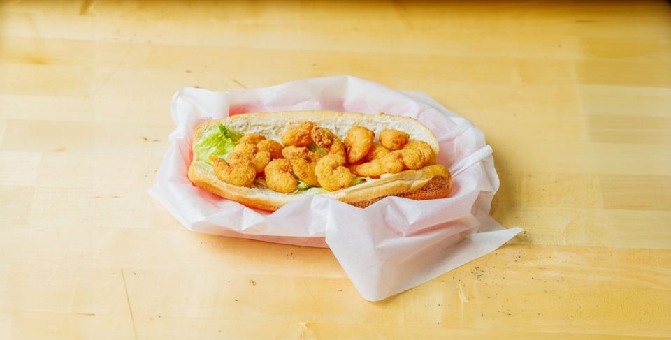 A shrimp po'boy sandwich with lettuce and sauce, wrapped in white paper on a wooden table.